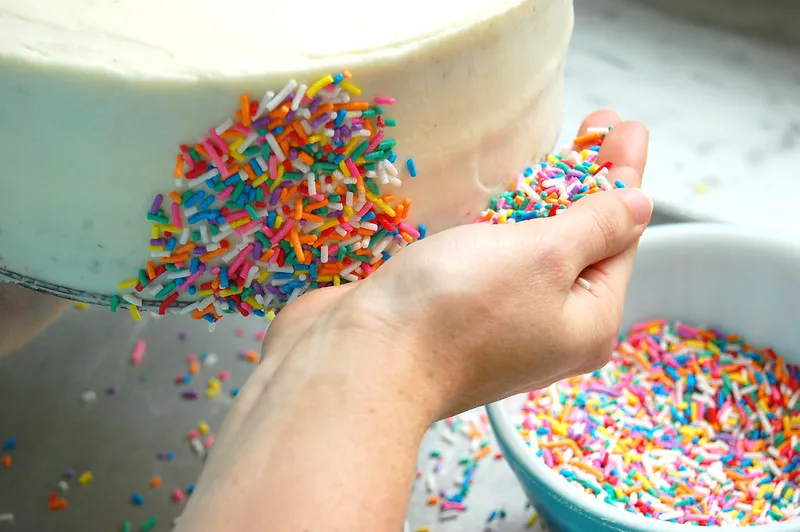 placing rainbow sprinkles on side of vanilla layer cake