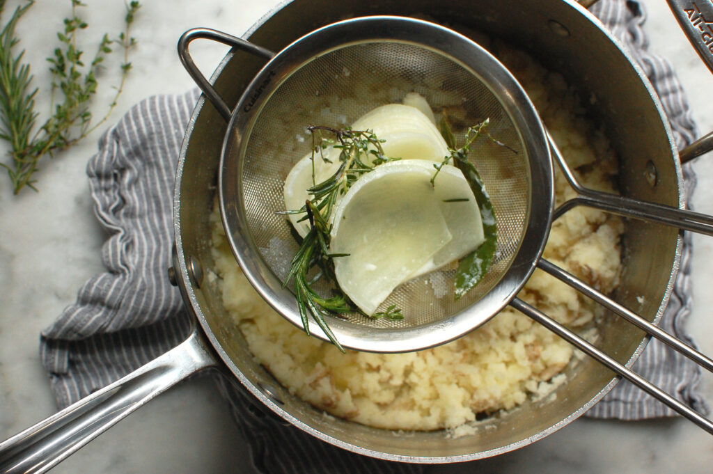 strainer of cooked aromatics over pot of mashed potatoes during cooking process