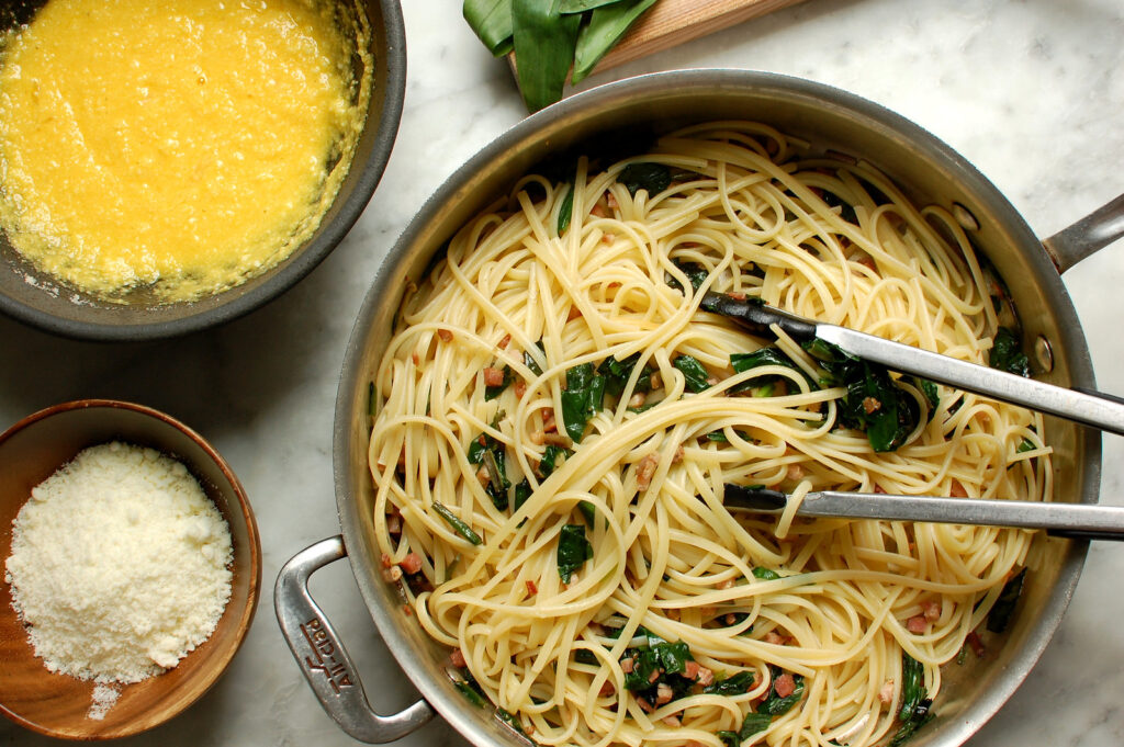 Spaghetti being tossed with pancetta and wild ramps in skillet before adding to egg and cheese carbonara sauce