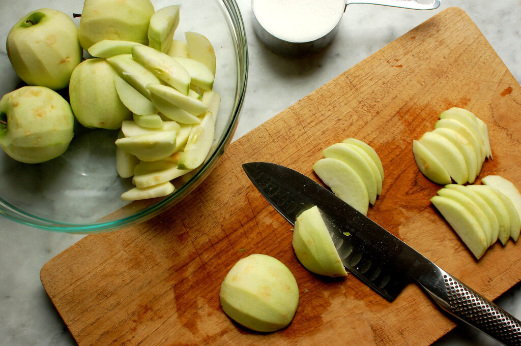 Peeled apples being sliced thin on cutting board before tossing with cinnamon sugar