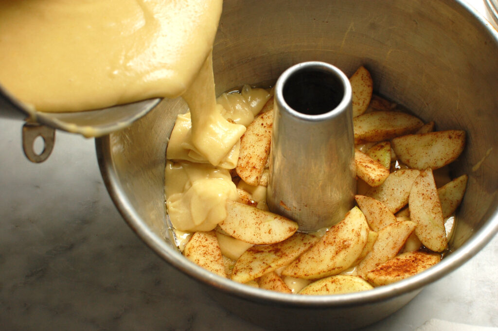 Thick, pale cake batter falling in a ribbon onto layer of apples in cake pan