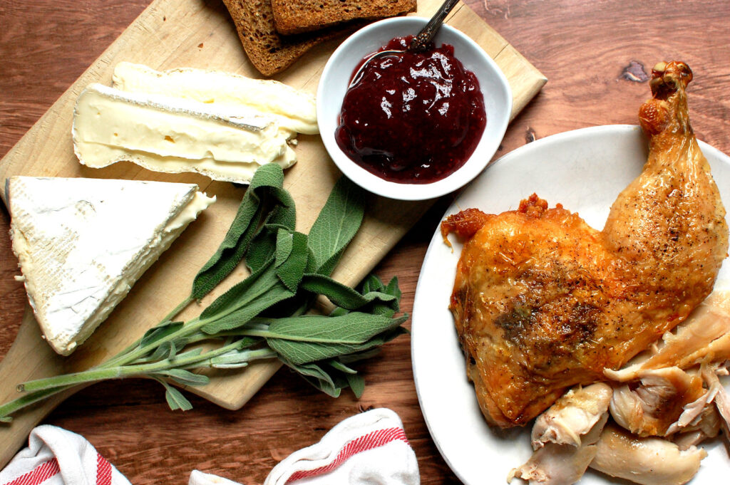 Styled shot of ingredients for a Cranberry Brie Turkey Melt: sliced roast turkey, a wedge of Brie, a bowl of cranberry sauce, and sliced wheat bread.