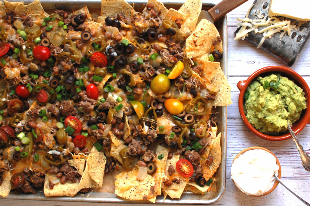 Oven baked sheet pan nachos loaded with seasoned ground beef, black beans, melted Monterey Jack cheese, guacamole, pico de gallo, and sliced scallions on a half-sheet pan