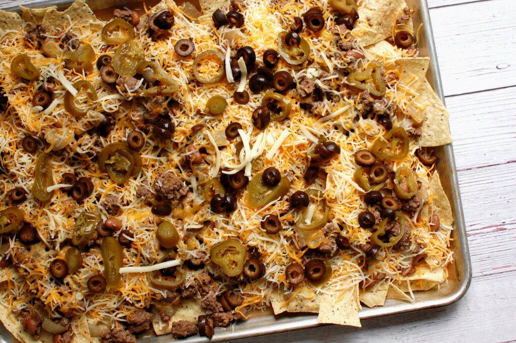 Sheet pan nachos fully assembled before baking: single layer of tortilla chips covered with seasoned ground beef, black beans, shredded Monterey Jack cheese before adding sliced pickled jalapeños, and drained black olives
