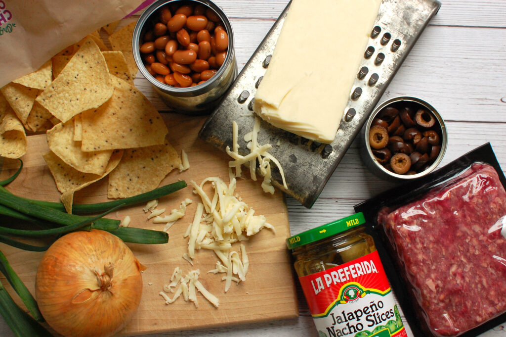Ingredients for oven baked nachos laid out on a surface: bag of tortilla chips, raw ground beef, can of black beans, shredded Monterey Jack cheese, pickled jalapeños, canned black olives, yellow onion, cumin, and kosher salt