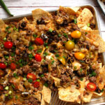 Oven baked sheet pan nachos loaded with seasoned ground beef, black beans, melted Monterey Jack cheese, guacamole, pico de gallo, and sliced scallions on a half-sheet pan