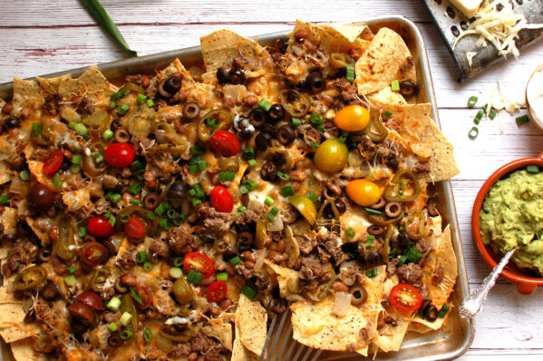 Oven baked sheet pan nachos loaded with seasoned ground beef, black beans, melted Monterey Jack cheese, guacamole, pico de gallo, and sliced scallions on a half-sheet pan