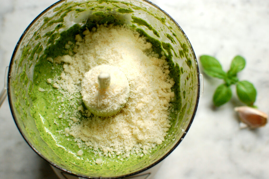 Blended basil pesto in bowl of food processor, green and silky, with grated cheese on top