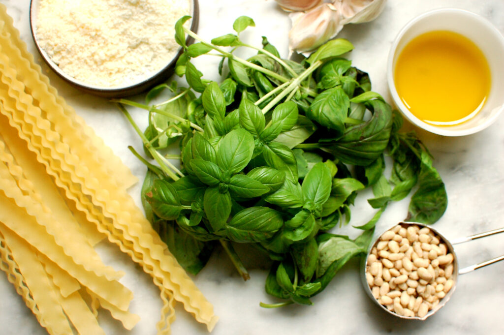 Overhead view of basil pesto ingredients: a bunch of fresh basil, pine nuts, garlic, two types of grated cheese, olive oil, and salt