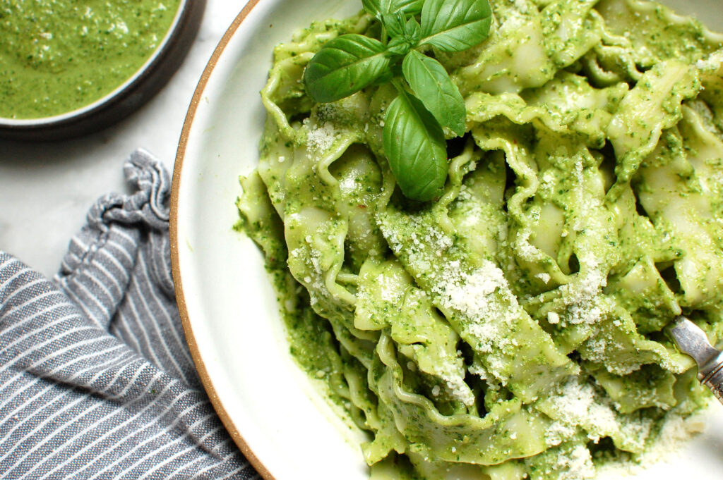 A white bowl of pappardelle basil pesto pasta tossed with bright green pesto, topped with grated Parmesan and a fresh basil sprig