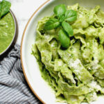 Bright green homemade basil pesto tossed with pasta in a white bowl with a spoon, surrounded by fresh basil leaves and bowl of silky green pesto