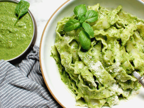 Bright green homemade basil pesto tossed with pasta in a white bowl with a spoon, surrounded by fresh basil leaves and bowl of silky green pesto