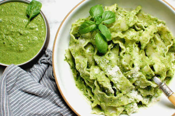 Bright green homemade basil pesto tossed with pasta in a white bowl with a spoon, surrounded by fresh basil leaves and bowl of silky green pesto