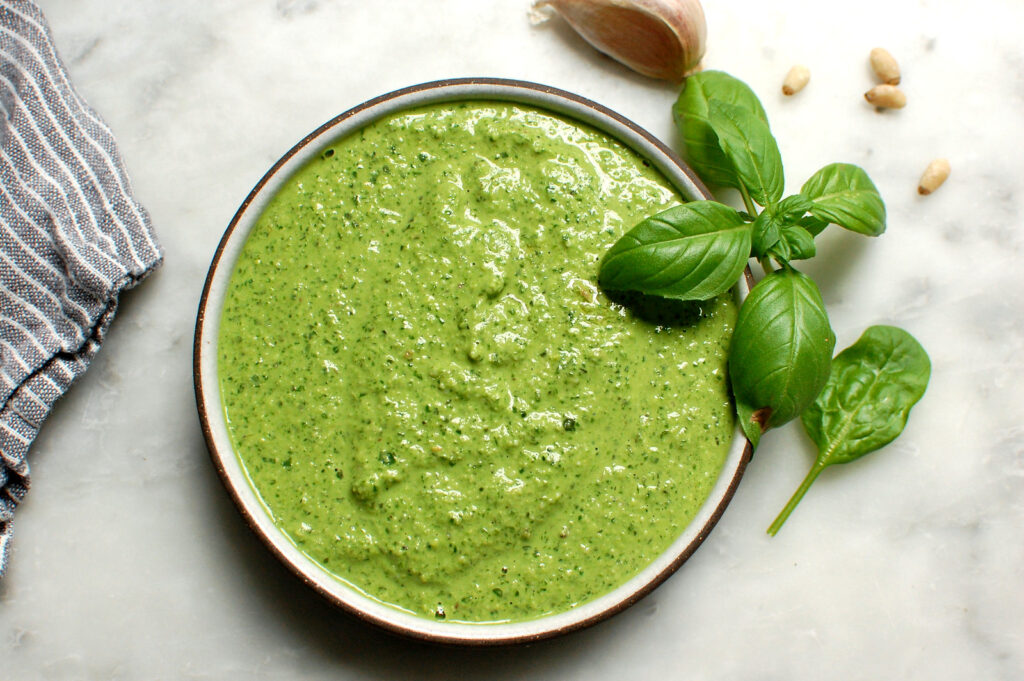 Bright green homemade basil pesto in a gray bowl with basil leaf garnish, surrounded by fresh garlic and pignoli.