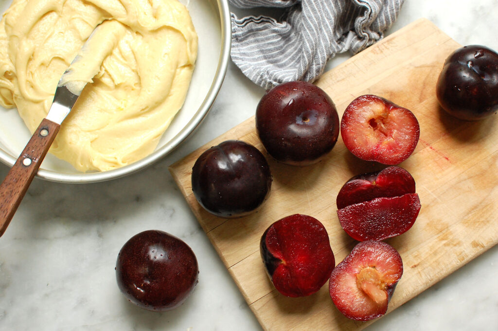 Recipe step of ripe plums being halved and pitted on cutting board before adding to cake batter in cake pan. 