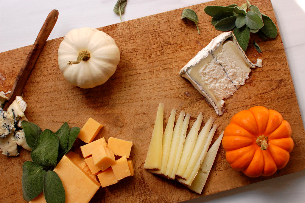 assembling autumn charcuterie board on wooden board with cheeses and fall decorations like herbs and mini pumpkins