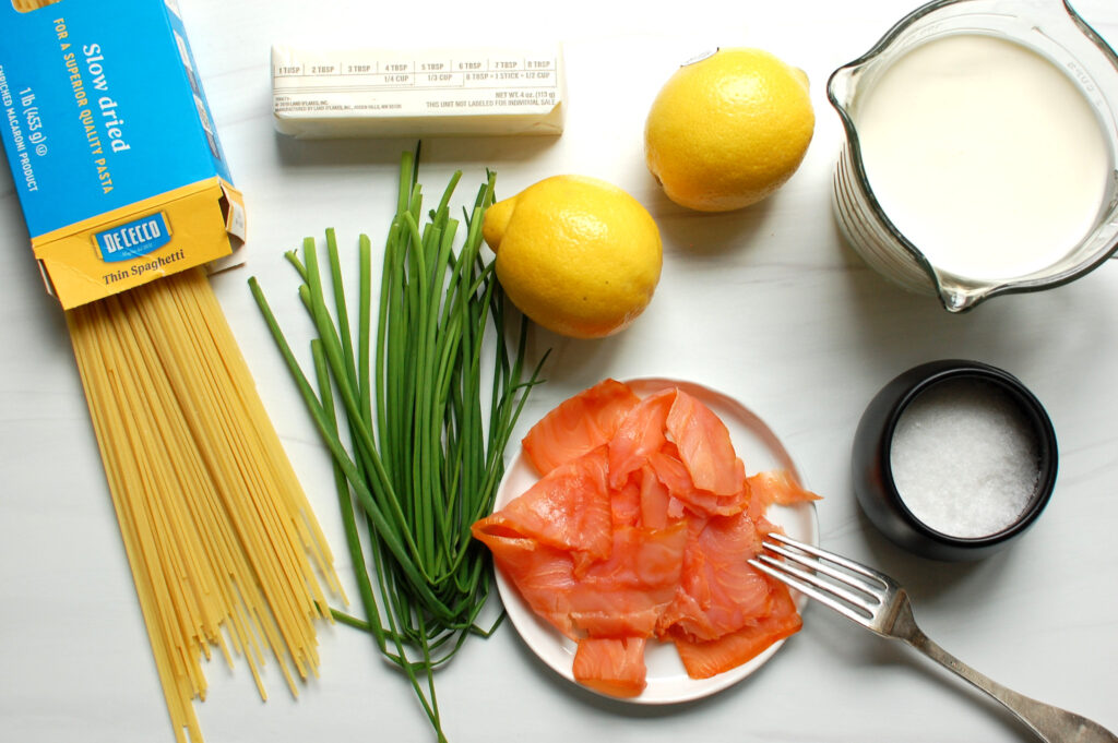 ingredients for creamy smoked salmon pasta on kitchen countertop, including smoked salmon, cream, lemon, pasta, butter, and chives