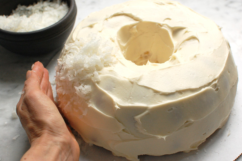 Applying shredded sweetened coconut to the bundt, which is frosted in cream cheese buttercream icing.