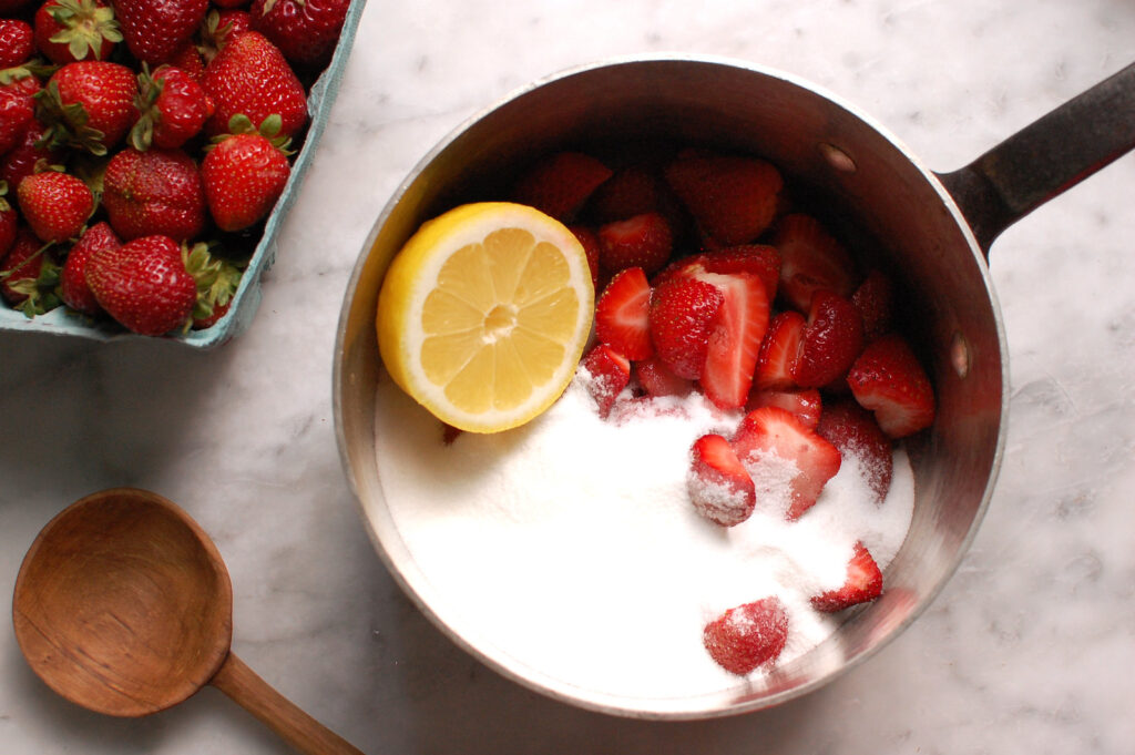 Chopped fresh strawberries combined with sugar and lemon juice in a saucepan to make strawberry preserves