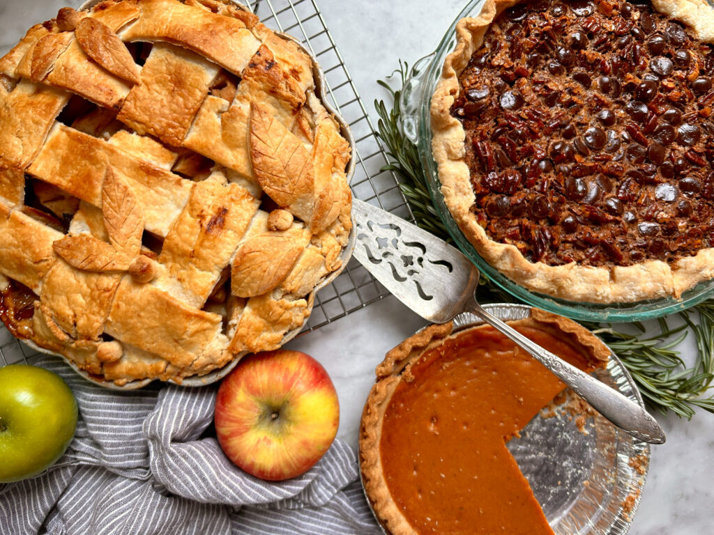 Warm overhead shot of a Thanksgiving dessert spread featuring apple pie, pumpkin pie, chocolate pecan pie, and pumpkin bread on a festive kitchen table