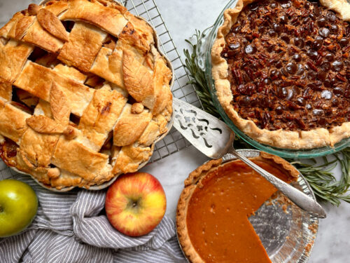 Warm overhead shot of a Thanksgiving dessert spread featuring apple pie, pumpkin pie, chocolate pecan pie, and pumpkin bread on a festive kitchen table
