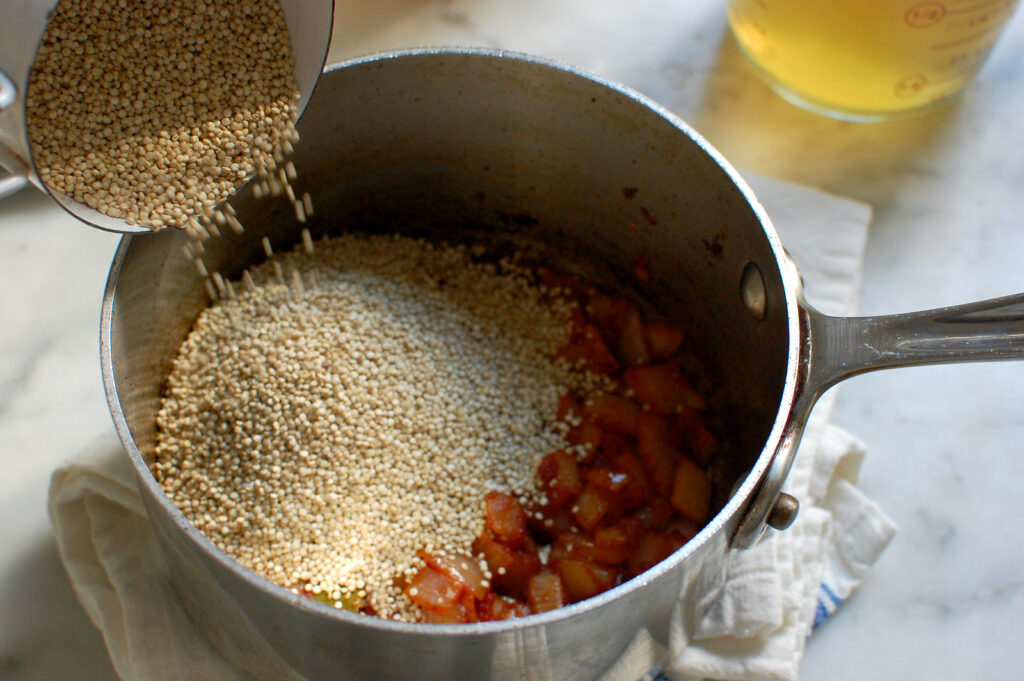 Adding dry quinoa to sautéed onions, garlic, and tomato paste in a saucepan.